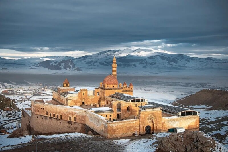 A large, historic stone palace with domed roofs and a tall minaret sits amid snowy mountains and a wide valley under a cloudy sky, illuminated by soft sunlight.