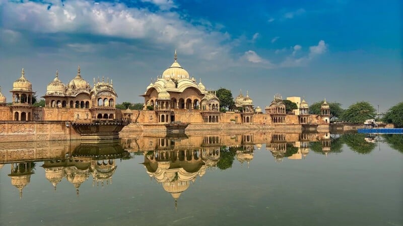 A row of ornate domed buildings with intricate carvings is reflected in a calm body of water under a blue sky with scattered clouds. Lush greenery surrounds the historic architecture.