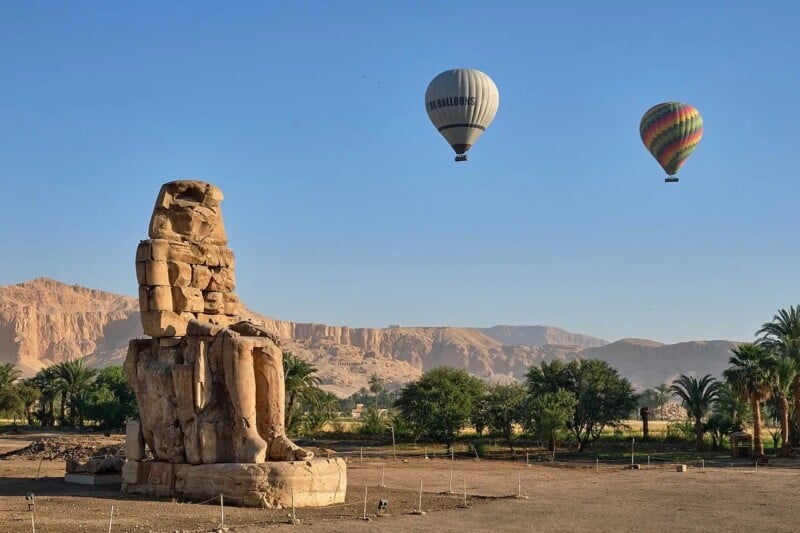 A large ancient stone statue sits in an open landscape with trees, mountains, and two colorful hot air balloons floating in the clear blue sky above.