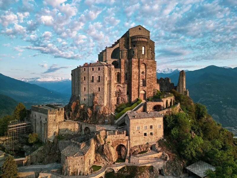 A large stone monastery sits atop a rocky hill surrounded by lush greenery, with distant mountains and a sky filled with scattered clouds in the background. The historic structure features arched windows and ancient walls.