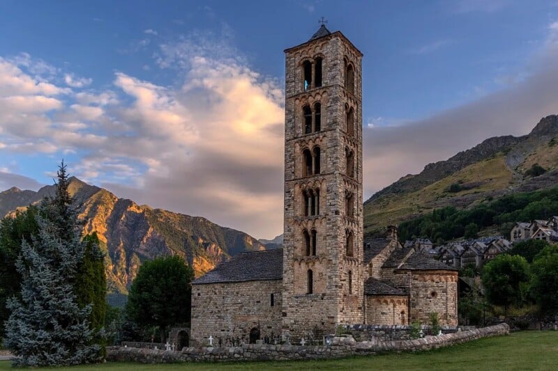 A stone church with a tall bell tower stands in a green landscape, surrounded by mountains and trees under a partly cloudy sky at sunset. The building features arched windows and rustic architecture.