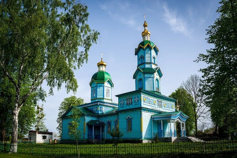 A vibrant blue and green wooden church with golden domes stands in a grassy area, surrounded by trees and a black metal fence under a clear blue sky.