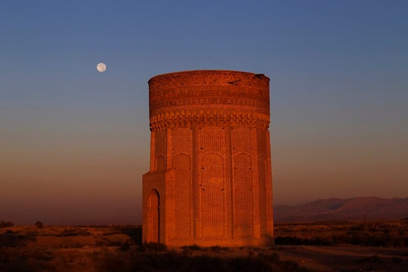 A round, brick tower with decorative patterns is illuminated by the warm light of sunset. The moon is visible in the clear sky, and mountains can be seen in the distance behind a desert landscape.