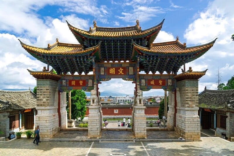 A traditional Chinese gate with ornate, multi-tiered roofs stands at the entrance to a scenic area, with people walking nearby and mountains visible in the background under a partly cloudy sky.
