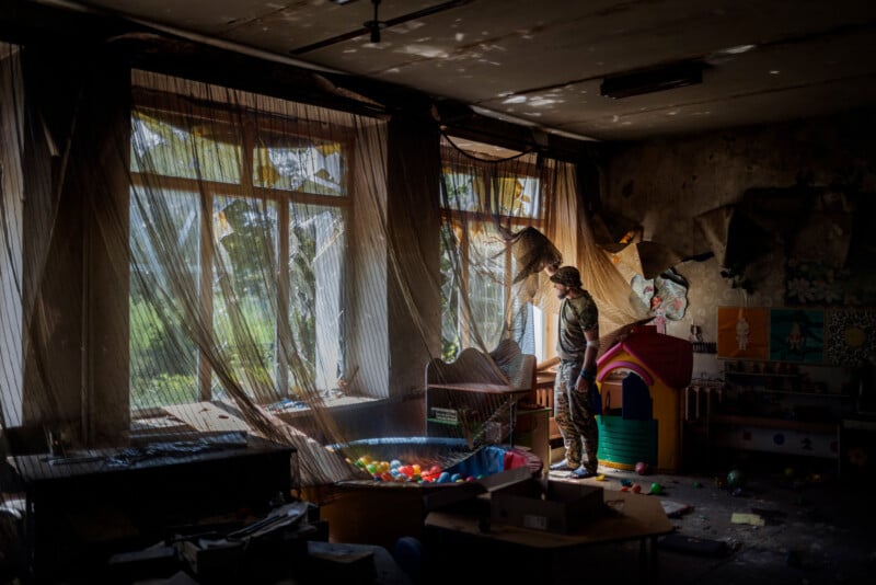 A soldier stands in a damaged, abandoned classroom filled with toys and a playhouse. Sunlight filters through torn curtains and broken windows, casting shadows across the debris-strewn room.