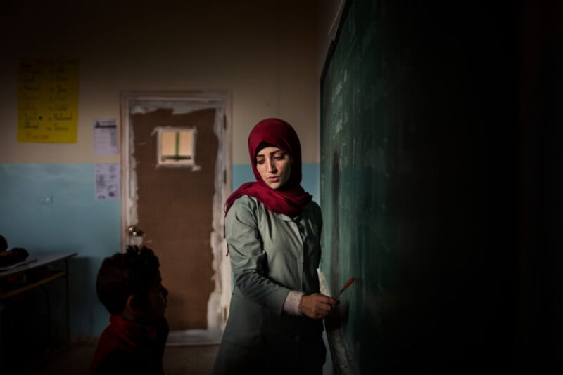 A woman wearing a red headscarf writes on a chalkboard in a dimly lit classroom, with a young student watching her. The background shows a door and some classroom posters.