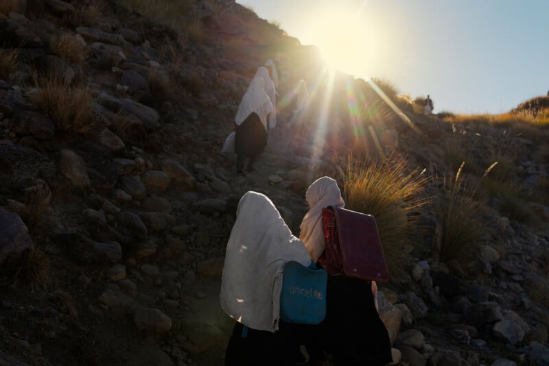 A group of girls wearing white headscarves and dark backpacks walk up a rocky hillside toward the sunlight, with rays of sun shining over the landscape.