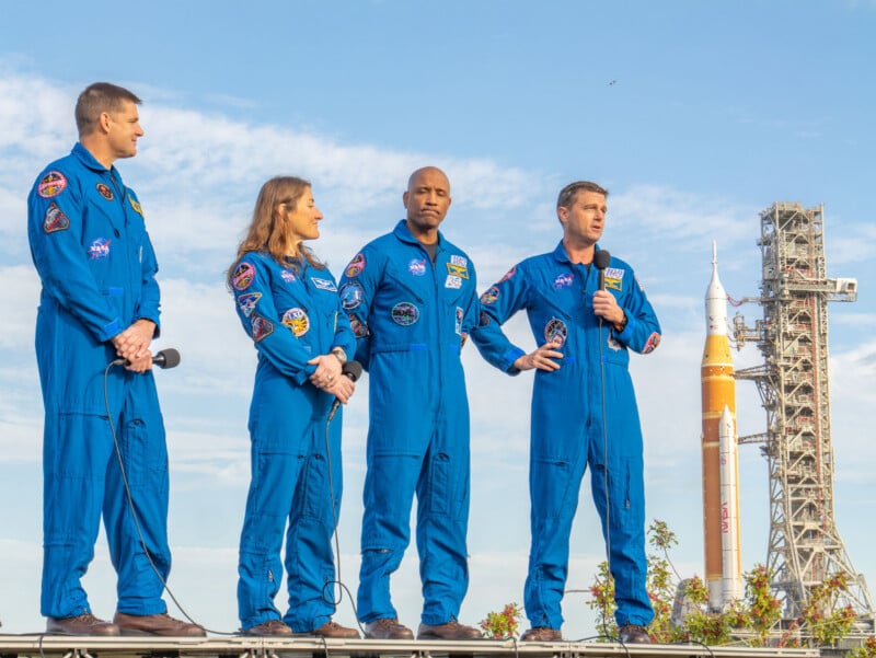 Four astronauts in blue NASA flight suits stand outdoors speaking in front of a launch pad with a large rocket in the background under a partly cloudy sky. One astronaut holds a microphone.