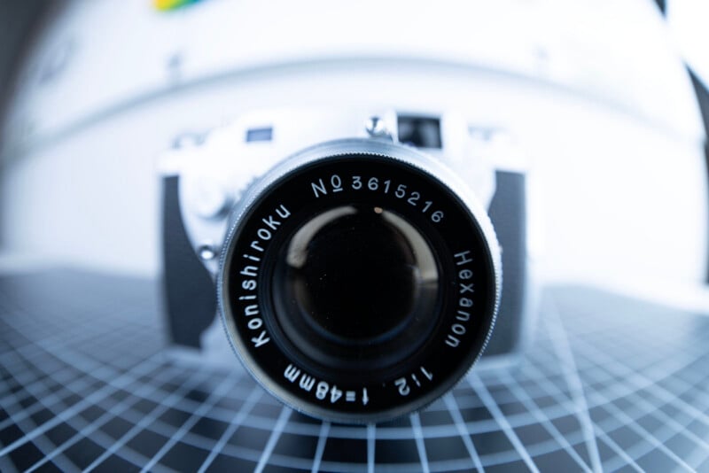 Close-up of a vintage Konishiroku Hexanon camera lens, focused on the lens markings, with a blurred white background and a black grid surface beneath the camera.