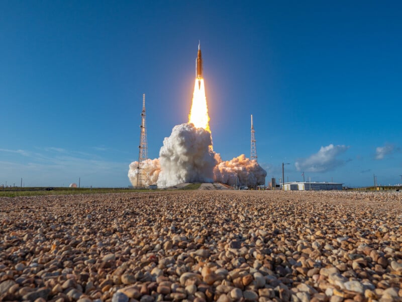 A rocket launches into a clear blue sky, emitting bright flames and thick smoke from its engines, with gravel in the foreground and launch towers on either side.