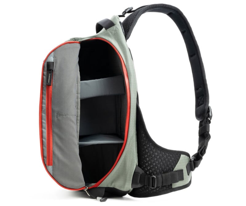 A gray sling backpack with a single padded strap, shown open to reveal a spacious main compartment with padded dividers. The interior is gray with a bright red zipper outlining the opening.