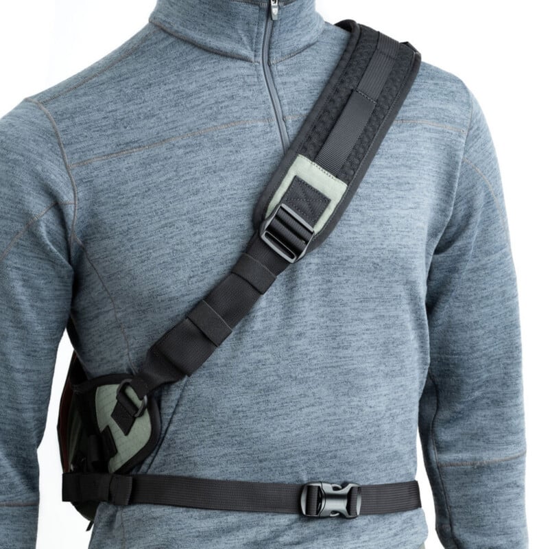 A person wearing a blue-gray zip-up pullover models a black crossbody strap harness with a buckle across the chest and waist, designed for carrying gear or equipment hands-free.