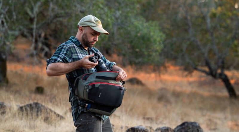 A man wearing a plaid shirt and cap stands outdoors in a dry, grassy area with trees, taking a camera out of a green and black bag slung across his shoulder.