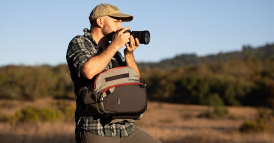 A person in a plaid shirt and cap stands outdoors in a field taking a photo with a camera, wearing a crossbody camera bag. Trees and hills are visible in the background under a clear sky.