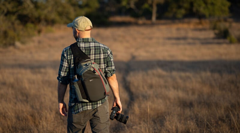 A person wearing a cap and plaid shirt walks through a dry grassy field with a camera bag slung over their shoulder and a camera in hand. Trees and sunlight are visible in the background.