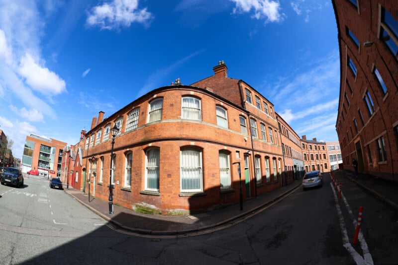A wide-angle view of a street corner with red-brick buildings under a bright blue sky with scattered clouds. Cars are parked along the street, and the buildings have many windows with white blinds.