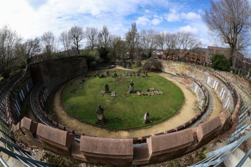 A wide-angle view of a circular, ancient structure with gravestones and grassy areas inside, surrounded by old stone walls and trees, under a partly cloudy sky.