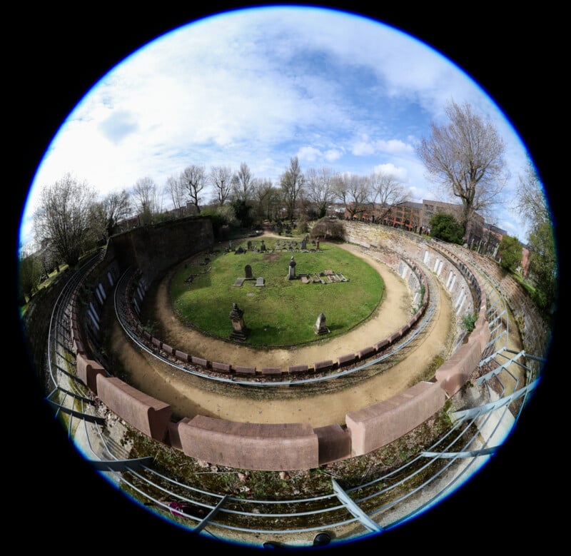 A fish-eye view of an ancient circular amphitheater with stone ruins and gravestones on grassy ground, surrounded by curved stone seating and leafless trees under a partly cloudy sky.