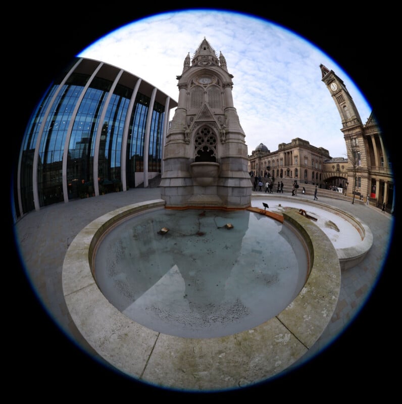 Fisheye view of a gothic-style clock monument with a shallow, circular reflecting pool in front, surrounded by historic and modern buildings under a partly cloudy sky.