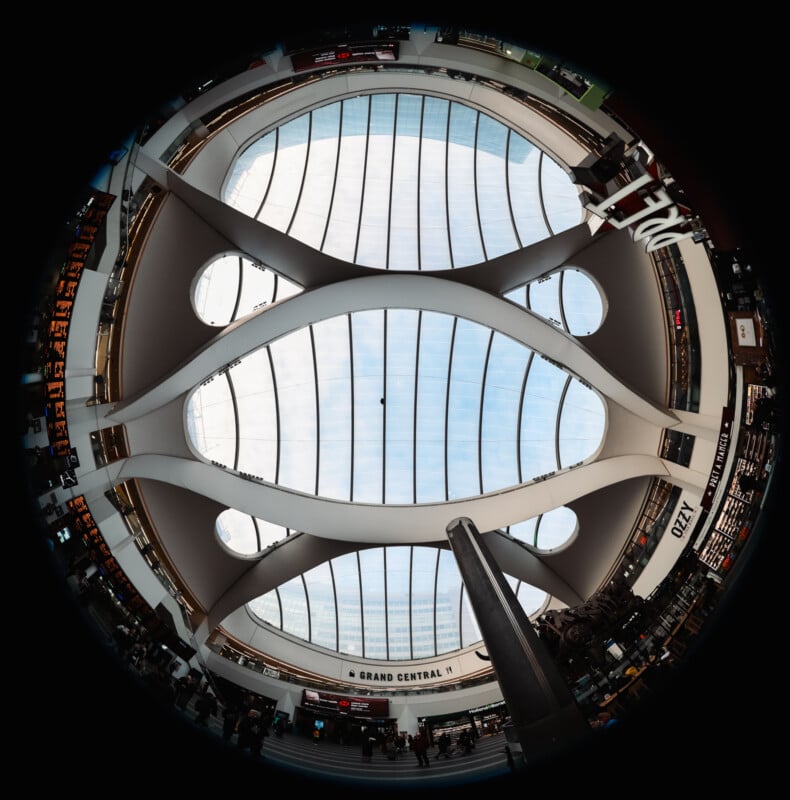 A fisheye view of a modern indoor space with a large, curved glass ceiling, intersecting arches, and multiple shops and signs, including one that reads "GRAND CENTRAL." Natural light floods the area.