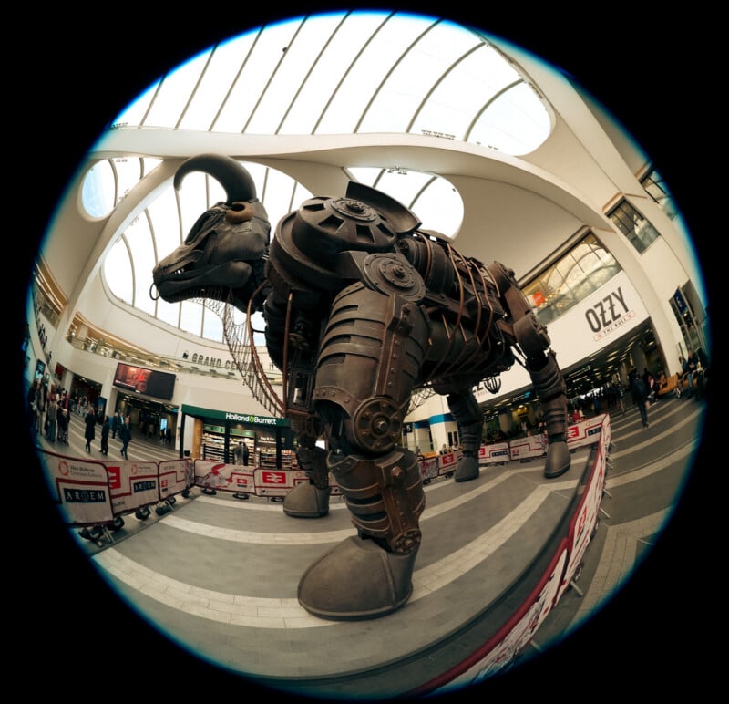 A large, steampunk-style robotic bull sculpture stands in a shopping mall, surrounded by safety barriers. The image is taken with a fisheye lens, creating a curved, wide-angle view of the indoor space.