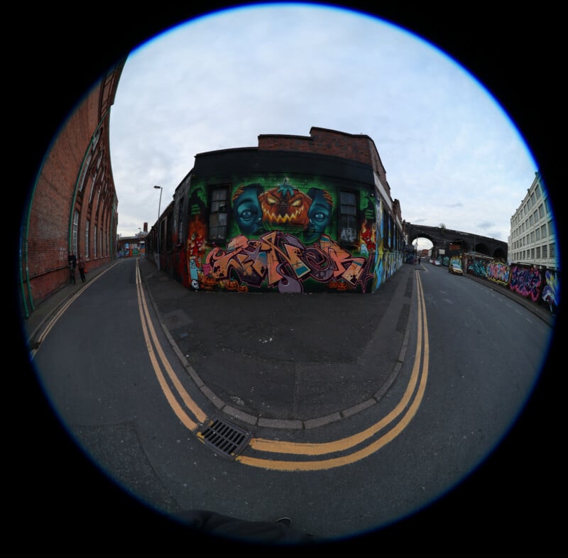 Fisheye view of a street corner with a brick building covered in colorful graffiti, including a large, green monster face. The road curves around the building, and the sky is overcast.