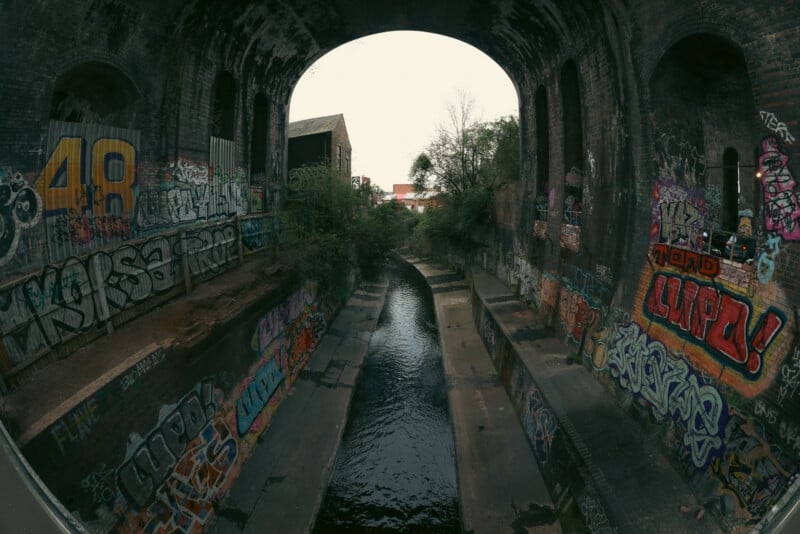A wide, arched brick tunnel with graffiti-covered walls and a narrow stream running through the center. Vegetation grows at the tunnel’s exit, and an overcast sky is visible outside.