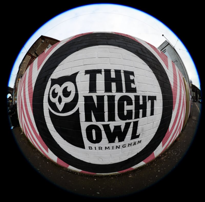 A fisheye lens photo of a mural featuring an owl face and the text "THE NIGHT OWL BIRMINGHAM" on a white, pink, and black circular background painted on a brick wall.