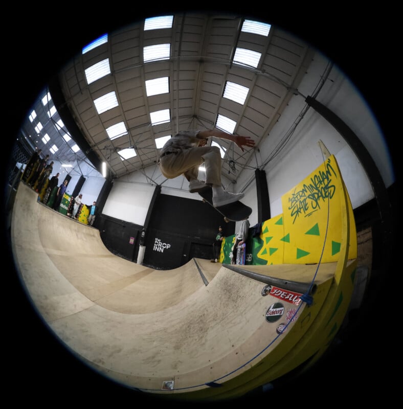 A skateboarder performs a trick mid-air above a ramp in an indoor skatepark, captured with a fisheye lens. The ramp is decorated with yellow and green graffiti and skateboard brand stickers.