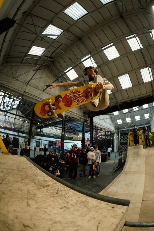 A skateboarder performs a high aerial trick indoors, holding his yellow skateboard decorated with cartoon graphics. Spectators watch from below in the industrial-style skatepark.