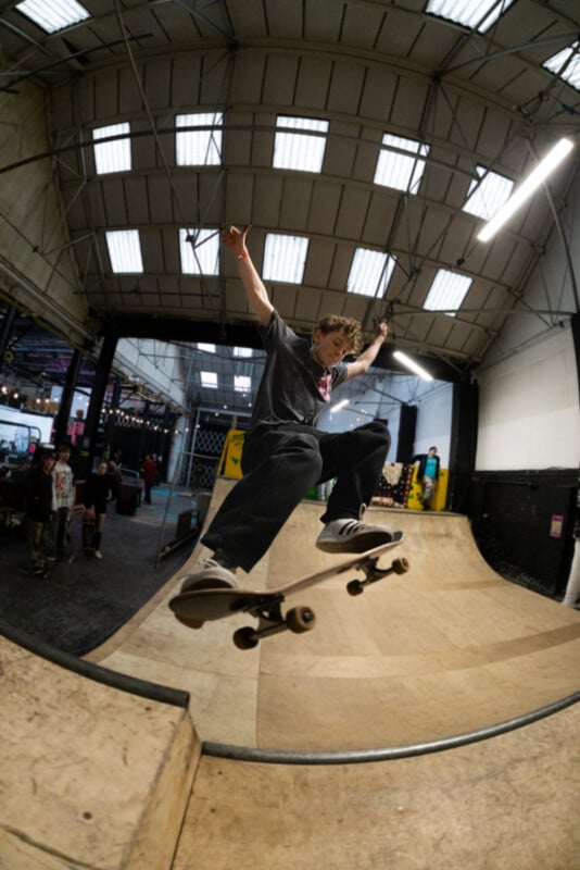 A skateboarder performs a trick in mid-air on an indoor skate ramp, with arms outstretched and knees bent, inside a warehouse-style skatepark under a high ceiling with skylights.