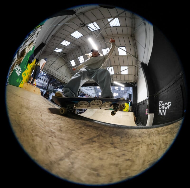 A fisheye view of a skateboarder grinding a ledge inside an indoor skatepark, with high ceilings, skylights, and graffiti on the walls. The scene is energetic and dynamic.