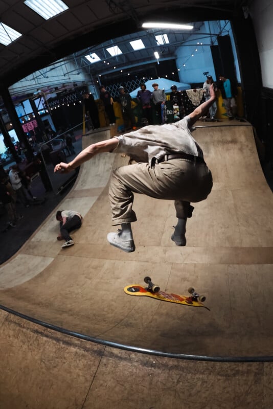 A skateboarder in mid-air performs a trick above a wooden ramp in an indoor skatepark, while other people watch and another skater sits on the ramp in the background.