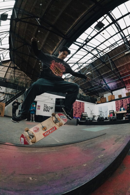 A skateboarder performs a trick indoors, flipping his board in mid-air. The background includes graffiti, a stage with musical equipment, and a few people watching in the skate park.
