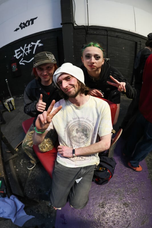 Three young adults pose indoors, all making peace signs. One sits on a red bench wearing a white beanie and tie-dye shirt; two stand behind, one with green hair and another in a red hat. "EXIT" is painted on the black wall.