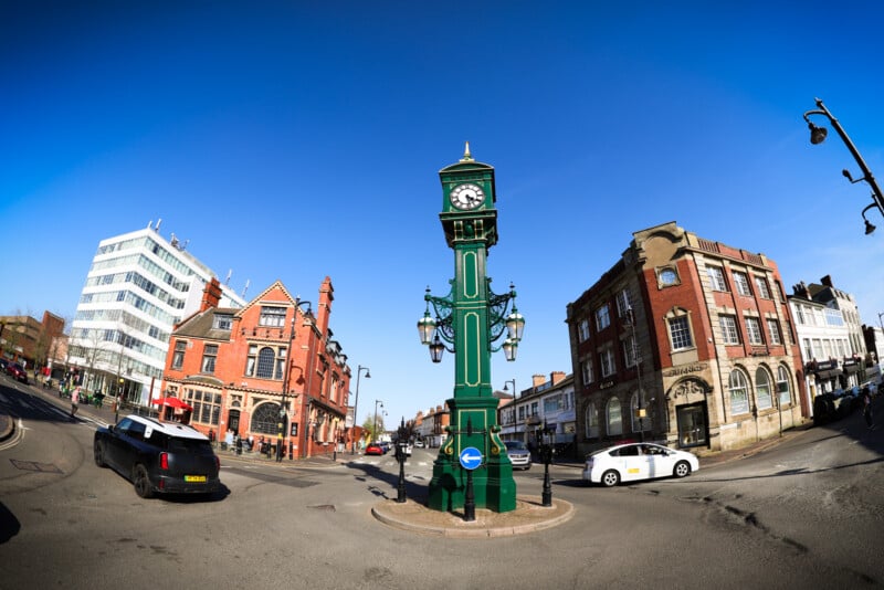 A green Victorian-style street clock stands at a road junction, surrounded by historic red-brick buildings, modern offices, and cars under a clear blue sky.
