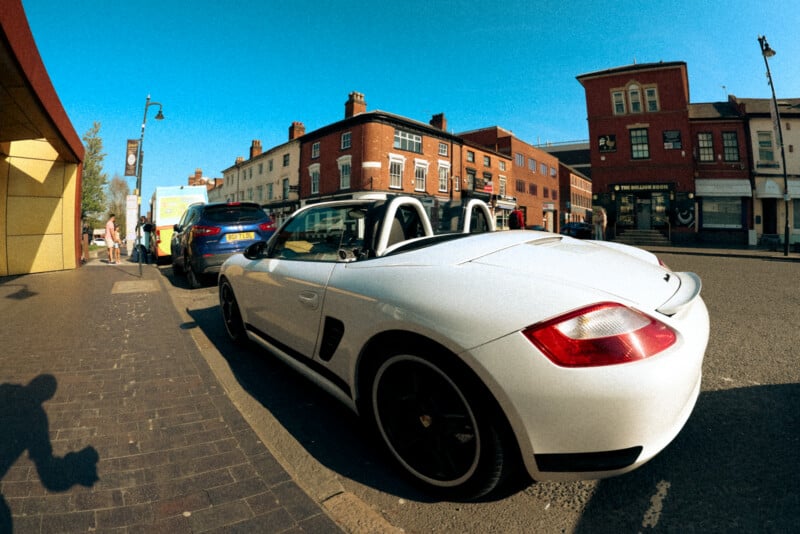 A white convertible sports car parked on a street in front of brick buildings under a clear blue sky, with other cars and pedestrians nearby.