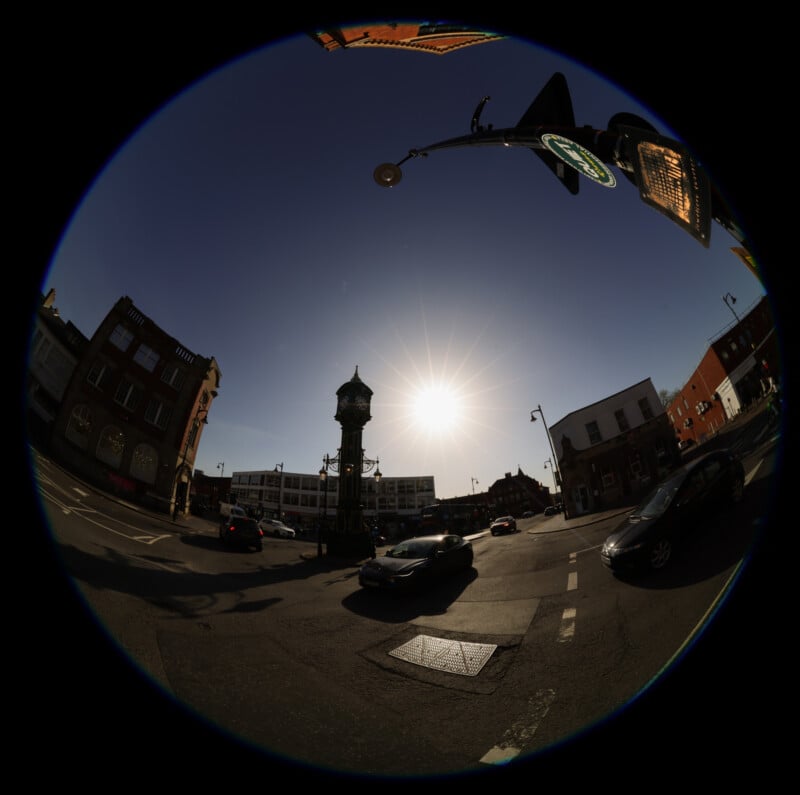 A fisheye lens view of a sunny urban street intersection with cars, old brick buildings, a streetlamp, and a central water tower, all under a clear blue sky with the sun shining brightly.