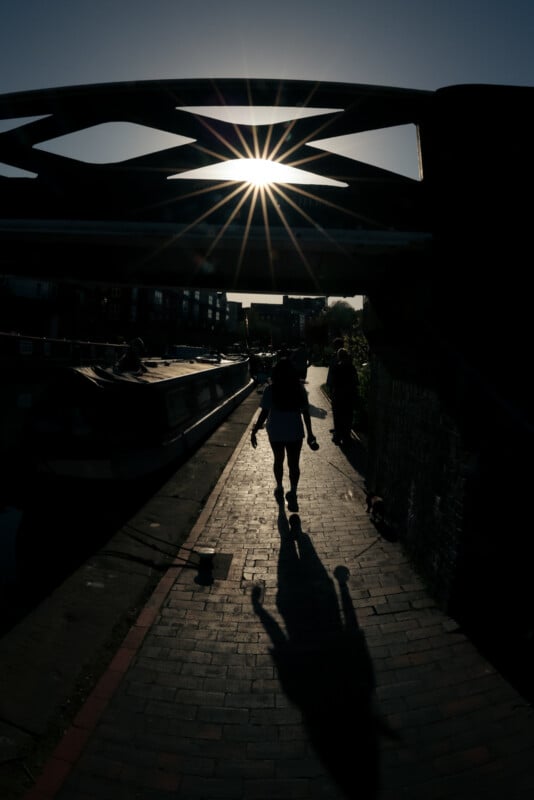 A person walks under a bridge beside a canal at sunset, casting a long shadow on the brick path as sunlight shines through the bridge’s geometric pattern.