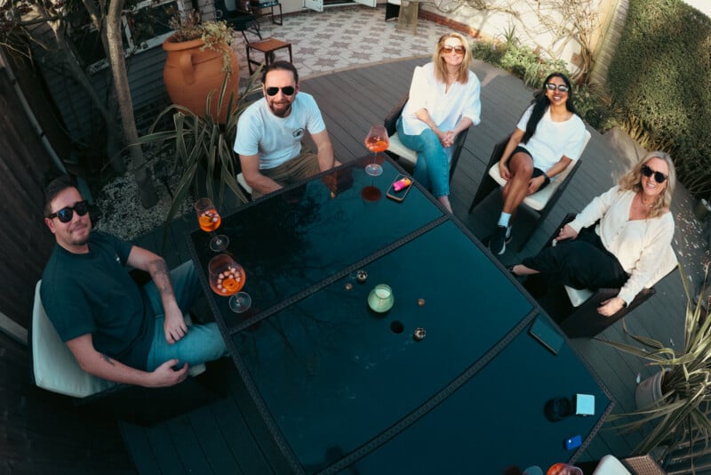 Five people sit around a glass outdoor table with drinks, smiling up at the camera on a sunny day. The scene is relaxed with plants and garden decor in the background.