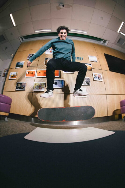 A man with curly hair, wearing a teal sweater and black pants, performs a trick on a skateboard indoors, mid-air, with a wall of photos and wood paneling in the background.