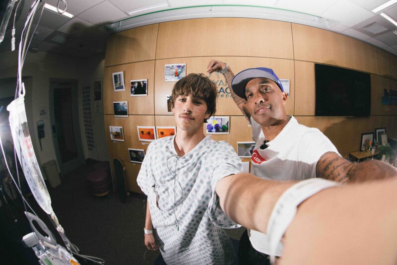 Two men pose for a selfie in a room with wood-paneled walls covered in photos. One wears a hospital gown and the other a cap and white shirt, extending his arm to take the picture. An IV stand is visible on the left.