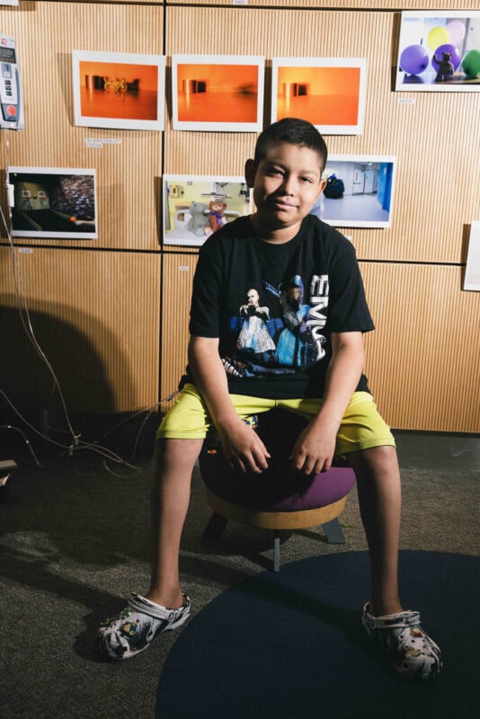 A boy with short hair sits on a purple stool, wearing a graphic T-shirt, yellow shorts, and patterned Crocs. Behind him, colorful photos are displayed on a wooden wall. The setting appears casual and artistic.