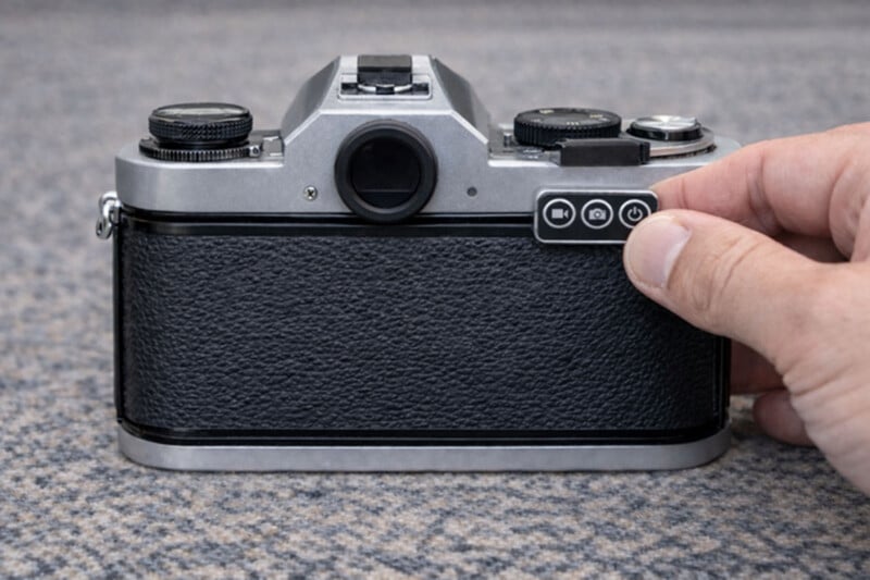 A hand holds a smartphone camera lens cap in front of the back of a vintage film camera, which is placed on a gray textured surface.