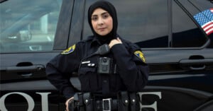 A police officer wearing a hijab stands confidently in front of a police vehicle, with an American flag decal visible on the car window.