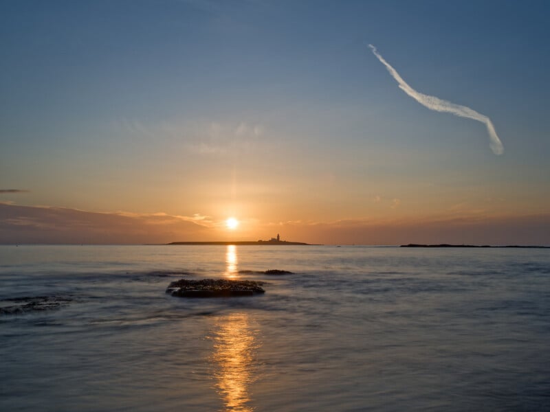 Calm sea at sunset with golden sunlight reflecting on the water, an island with a lighthouse on the horizon, and a streak of cloud in the blue sky.