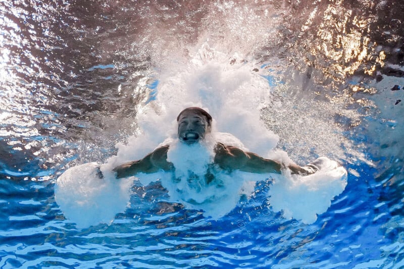 A swimmer captured underwater right after diving in, arms outstretched and surrounded by bubbles, with blue water and light reflections visible above.