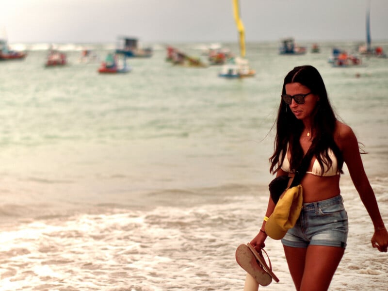 A woman in sunglasses, a white bikini top, and denim shorts walks along the shore, holding sandals, with boats floating on the sea in the background.