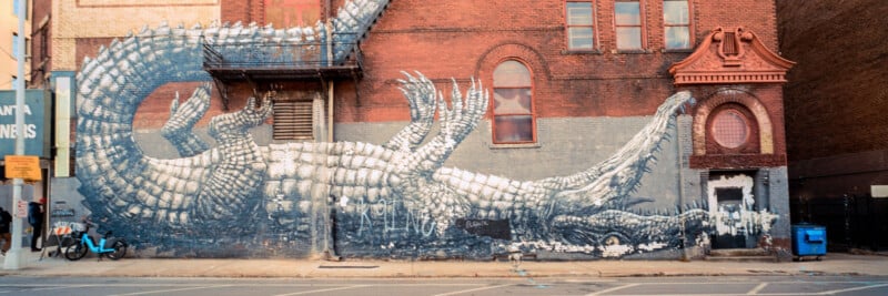 A large mural of a white and gray crocodile stretches across the red brick wall of a city building, wrapping around windows and doors on an urban street.