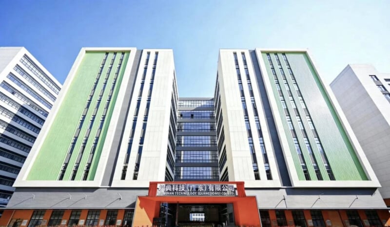 A modern multi-story office building with green vertical panels, large windows, and a central entrance. A red sign with Chinese text is displayed above the entrance. The sky is clear and blue.